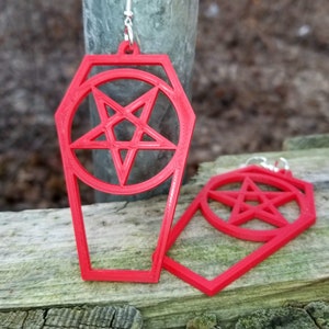 May include: A pair of red coffin-shaped earrings with a pentagram inside each. The earrings are made of plastic and have silver hooks.