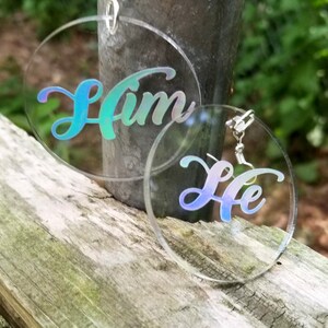 May include: A pair of iridescent clear acrylic earrings with the words "Him" and "Her" written in a cursive font.