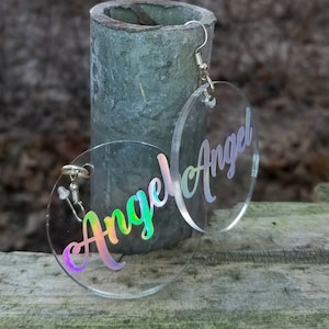 May include: A pair of iridescent clear acrylic earrings with the word "Angel" written in a cursive font.