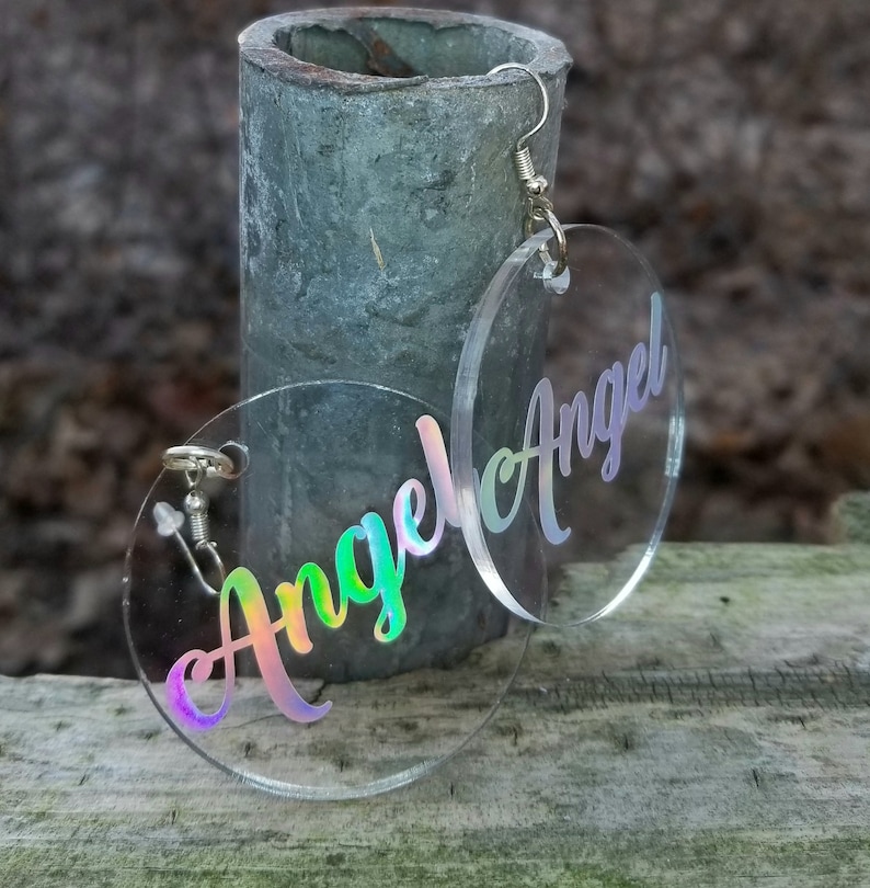 May include: A pair of iridescent clear acrylic earrings with the word "Angel" written in a cursive font.