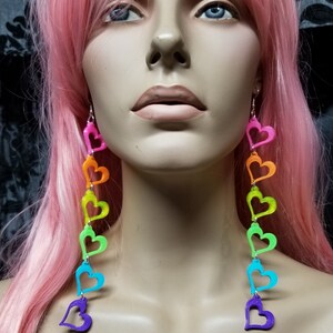 May include: Rainbow heart dangle earrings with a silver hook. The hearts are layered and are in the colors of the rainbow, starting with pink and ending with purple.