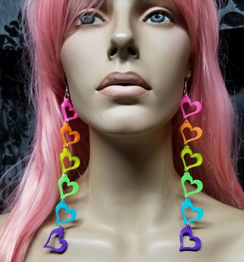 May include: Rainbow heart dangle earrings with a silver hook. The hearts are layered and are in the colors of the rainbow, starting with pink and ending with purple.