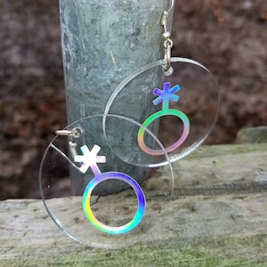 May include: A pair of iridescent clear acrylic earrings with a gender symbol design. The symbol is a circle with a cross on top and a smaller circle inside the larger circle. The earrings are hanging from silver hooks.