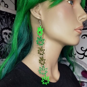Peut inclure: Une paire de boucles d'oreilles longues et pendantes avec une chaîne de breloques en forme de feuilles de marijuana vertes et dorées.