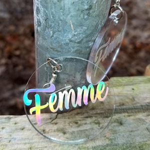 May include: A pair of iridescent clear acrylic earrings with the word "Femme" written in a rainbow script.