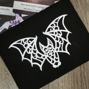 May include: A white, intricately designed bat silhouette on a black background. The bat has a detailed, lacy pattern on its wings and body.