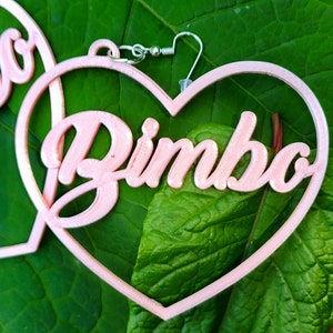 K&ouml;nnte beinhalten: Rosa herzf&ouml;rmige Ohrringe mit dem Wort "Bimbo" in 3D-Optik.