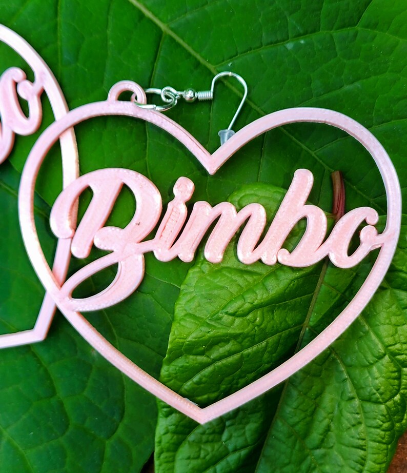 K&ouml;nnte beinhalten: Rosa herzf&ouml;rmige Ohrringe mit dem Wort "Bimbo" in 3D-Optik.