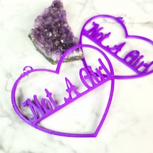 May include: A pair of purple heart-shaped earrings with the words "Not a Girl" cut out in the center of each heart.