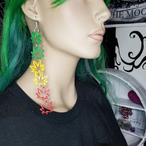 May include: A pair of long, dangling earrings featuring a cascade of green, yellow, and red marijuana leaf charms.