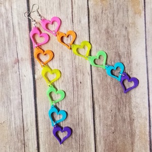 May include: Rainbow heart shaped earrings with a silver hook. The hearts are connected in a chain and are in the colors red, orange, yellow, green, blue, indigo, and violet.