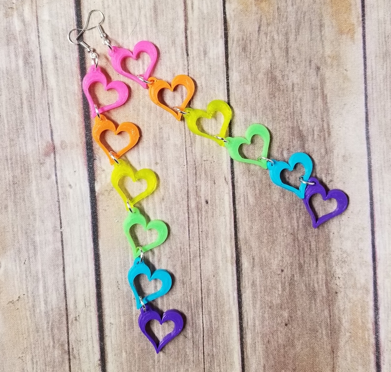 May include: Rainbow heart shaped earrings with a silver hook. The hearts are connected in a chain and are in the colors red, orange, yellow, green, blue, indigo, and violet.