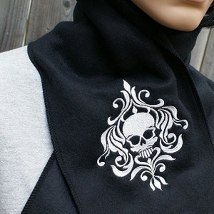 Puede incluir: Cuello de forro polar negro con un dise&ntilde;o de calavera y flores bordadas en blanco.