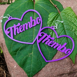 May include: A pair of purple heart-shaped earrings with the name "Thembo" in a cursive font. The earrings are displayed on a large green leaf, with a brown and green background.