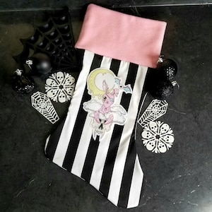 May include: A black and white striped Christmas stocking with a pink cuff. The stocking features a pink and white cartoon character with a single eye and bat wings, sitting on a cloud with a crescent moon and dripping yellow liquid.