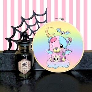 May include: A pastel rainbow gradient embroidery hoop with a stitched image of a pink and blue patchwork teddy bear with devil wings and a skull. The teddy bear has a stitched X on its forehead and a dripping yellow substance on its face. The background includes a crescent moon and a bat with an eye.