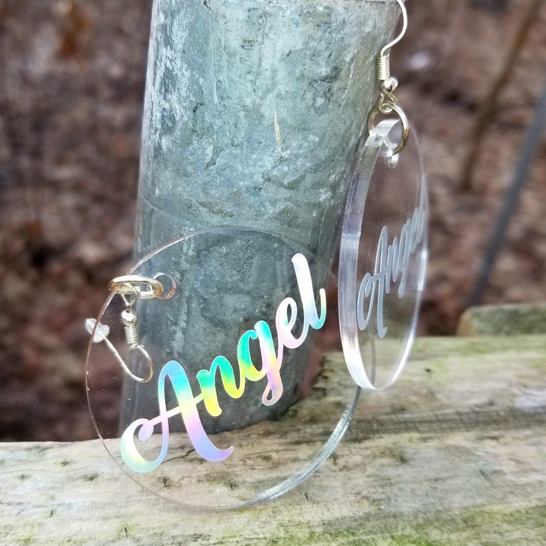 May include: A pair of clear acrylic earrings with the word "Angel" written in iridescent holographic lettering.