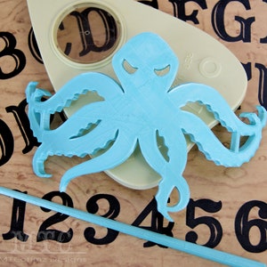 May include: A light blue octopus cutout with a menacing expression. The octopus is placed on a wooden surface with black numbers and letters.
