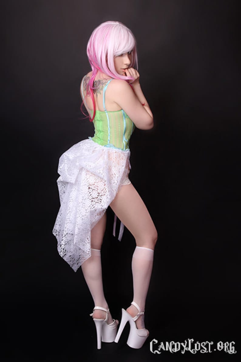 Pu&ograve; includere: Una donna indossa una gonna di pizzo bianco con un corpetto a righe verdi e bianche, una parrucca rosa e tacchi bianchi.