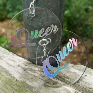 May include: A pair of iridescent clear acrylic hoop earrings with the word "Queer" written in a cursive font on each earring.