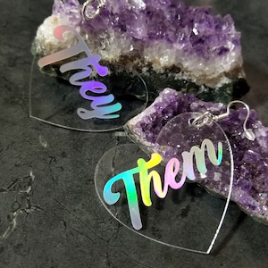 May include: A pair of iridescent heart-shaped earrings with the words "they" and "them" written on them. The earrings are hanging from a purple amethyst crystal.