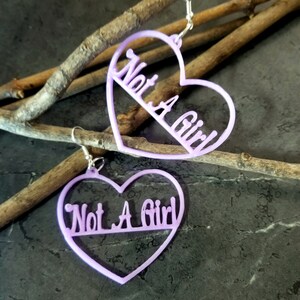 May include: A pair of lavender heart-shaped earrings with the text "Not A Girl" written inside each heart.