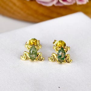 May include: A pair of gold-colored frog stud earrings. Each earring features a frog design with a green, oval-shaped stone body. The earrings are set on a white surface, with a blurred floral arrangement in the background.