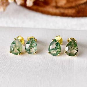 May include: Three pairs of gold-colored stud earrings with teardrop-shaped, moss agate stones. The stones have a translucent base with green, fern-like inclusions. The earrings are displayed on a white surface with a blurred, natural background.