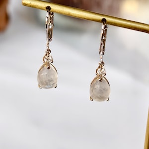 Puede incluir: Un par de pendientes dorados con una piedra translúcida en forma de lágrima. Cada pendiente tiene una pequeña gema transparente en la parte superior y un cierre de palanca. Los pendientes se exhiben en un expositor de joyería dorado.