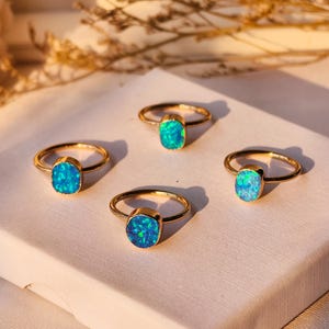 May include: Four gold rings with vibrant blue opal stones. The rings are arranged on a white surface, with a soft, warm light illuminating them. The opal stones have a captivating, iridescent quality.