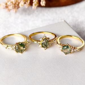 Può includere: Tre anelli d'oro con pietre preziose verdi e bianche. Gli anelli presentano diversi design, tra cui una pietra centrale circondata da gemme più piccole e un motivo a stella. Sono esposti su una superficie bianca.