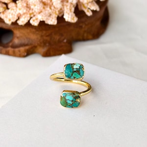 May include: A gold ring featuring two rectangular turquoise stones. The ring's band has a distinctive, open design that wraps around the finger. The turquoise stones display a blend of blue and brown hues.