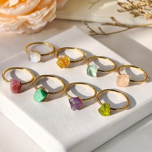 May include: A collection of gold-toned rings, each featuring a unique, raw gemstone. The gemstones vary in color, including pink, green, purple, yellow, and clear. The rings are displayed on a white surface, with soft floral accents in the background.
