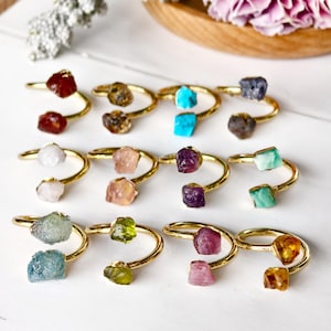 Può includere: Una collezione di anelli regolabili color oro, ognuno con una gemma grezza unica. Le gemme variano di colore, tra cui rosso, marrone, turchese, viola, verde e rosa. Gli anelli sono esposti su una superficie bianca.