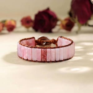 May include: Pink gemstone bracelet with a brown leather band and a silver clasp. The bracelet features rectangular pink stones. The leather band is stitched around the stones. The bracelet is a wrap-around style.