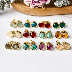 Op de afbeelding: Een verzameling goudkleurige oorstekers met diverse kleurrijke edelstenen. De oorbellen hebben ronde en facetgeslepen stenen in groen, blauw, rood, geel en roze tinten. De oorbellen worden gepresenteerd op een wit oppervlak.