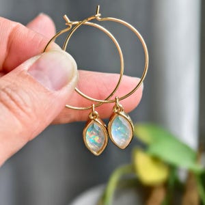 Könnte beinhalten: Goldene Creolen mit tropfenförmigen, schillernden Anhängern. Die Anhänger haben einen Regenbogen-Schimmer, und die Creolen werden von einer Hand gehalten. Die Ohrringe sind ein Schmuckstück.