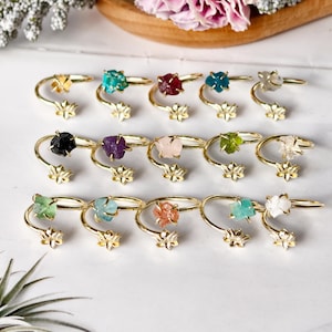 Op de afbeelding: Een verzameling goudkleurige ringen, elk met een unieke, onregelmatig gevormde edelsteen in verschillende kleuren. De edelstenen omvatten tinten groen, blauw, rood en paars. De ringen hebben een lotusbloem-ontwerp.