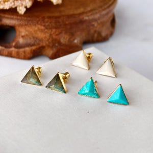 Puede incluir: Seis pendientes de botón dorados con diseños triangulares geométricos. Los pendientes presentan tres colores diferentes: turquesa, crema y un verde oscuro iridiscente. Los pendientes se muestran sobre una superficie blanca.
