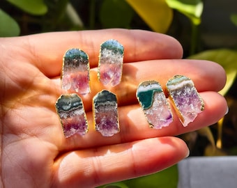 May include: A collection of six raw gemstone earrings with gold-colored edges. The gemstones display a mix of colors, including pink, purple, green, and blue. The earrings are displayed on a hand, showcasing their natural beauty and unique shapes.