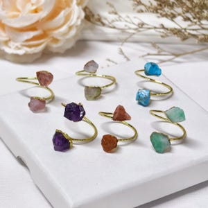 May include: A collection of gemstone rings with a spiral design. Each ring features a gold-colored band and two raw, uncut gemstones in various colors, including pink, purple, blue, and green. The rings are displayed on a white surface.