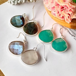 May include: A collection of gemstone earrings with silver-colored hooks. The earrings feature teardrop-shaped stones in various colors, including blue, green, and white. The stones are set in a silver bezel. The earrings are displayed on a white surface.