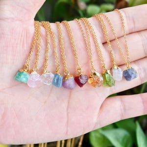 Peut inclure: Plusieurs colliers dorés avec des pendentifs en pierres brutes de différentes couleurs : vert, rose, transparent, bleu, violet, rouge, orange et gris. Les colliers sont présentés sur une main.