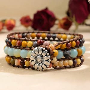 Könnte beinhalten: Mehrsträngiges Wickelarmband mit silbernem Blumenverschluss. Das Armband besteht aus braunem Leder und farbigen Edelsteinperlen in Gelb-, Blau-, Braun- und Rosatönen. Das Armband ist im Bohemian-Stil gehalten.