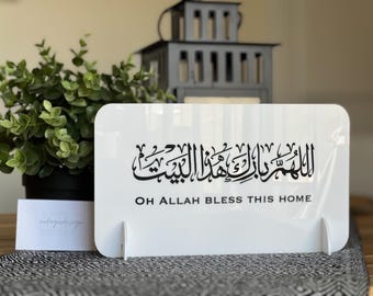 Op de afbeelding: Een wit acrylen decoratiebord met zwarte Arabische kalligrafie en de Engelse tekst "OH ALLAH BLESS THIS HOME". Het bord wordt ondersteund door twee witte steunen. Een potplant en een decoratieve lantaarn staan op de achtergrond.
