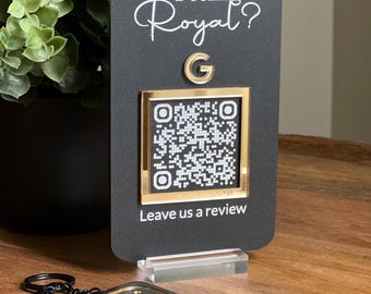 May include: Black sign with gold trim featuring a QR code and the text "Feeling Royal?" with a gold letter G. The sign is on a stand and a black keychain with a gold ring is in the foreground.