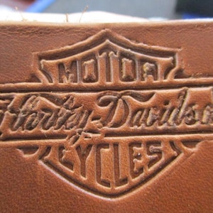 Puede incluir: Un parche de cuero marrón con el logotipo de Harley-Davidson en relieve en forma de escudo. El logotipo presenta las palabras "Motor" en la parte superior, "Harley-Davidson" en una fuente de escritura en el centro y "Cycles" en la parte inferior.