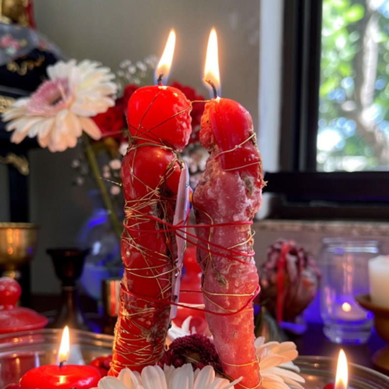 May include: Two red, figure-shaped candles with lit wicks, bound together with gold wire and red string. The candles are surrounded by white and red flowers, with a blurred background of a window and other candles.