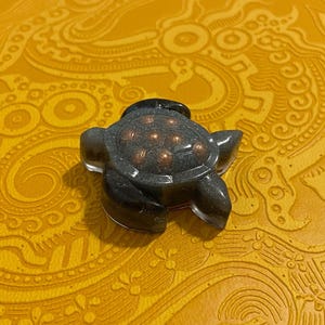 May include: A small, dark gray turtle figurine with a textured shell and visible copper-colored inclusions. The turtle is set against a yellow background with an ornate pattern. The figurine is a decorative item.