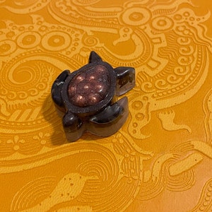 May include: A small black resin turtle figurine with a copper-colored shell and a glittery finish. The turtle is sitting on a yellow surface with a decorative pattern.